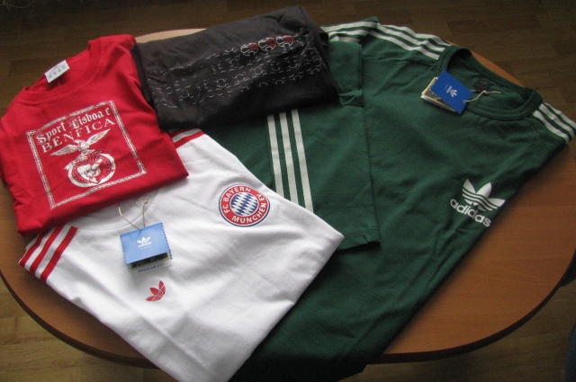 Stos ubrań sportowych na okrągłym stole: czerwona koszulka Sport Lisboa e Benfica, biała koszulka Bayern Monachium, czarna koszulka z wzorem i zielona koszulka Adidas z trzema białymi paskami...